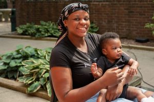 A mother holding her infant son. The mother is smiling at the camera, the infant is staring into the distance