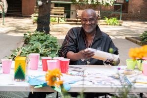 An individual sitting at an art activity table, holding an art canvas, smiling at the camera