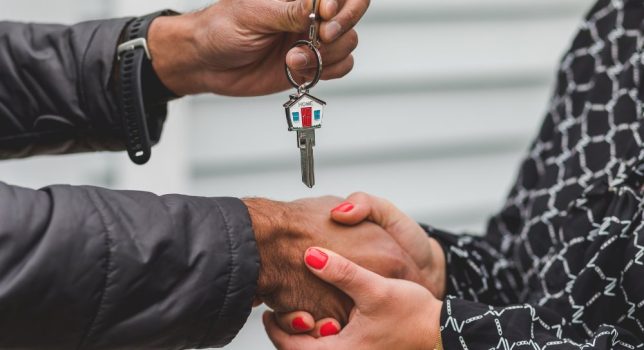Two individuals shaking hands, one of the individuals holding up a key with a house painted on it.